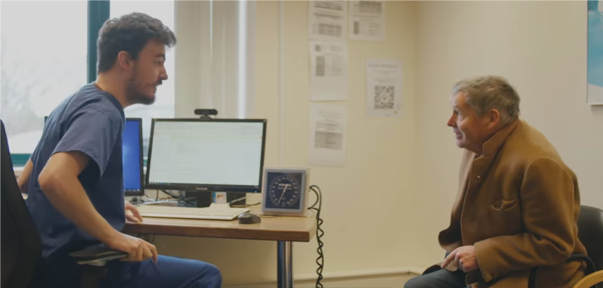 A healthcare assistant speaking with an older man in a GP surgery clinic room